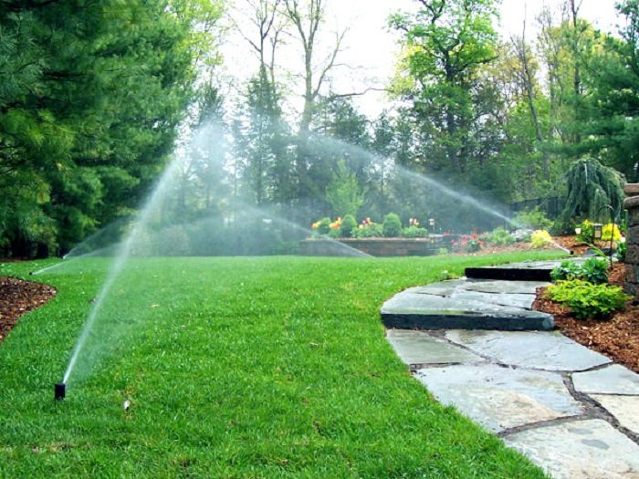 Tưới Cây Tự Động – Cam Kết Một Khu Vườn Luôn Xanh Dù Bạn Bận Rộn 3 Hệ thống tưới cây tự động đang phun nước đều trên thảm cỏ trong một sân vườn nhỏ, kết hợp đầu tưới âm sàn và cảnh quan xanh mát.